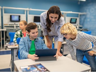 Учителка показва на своите ученици как да боравят с електронно устройство.